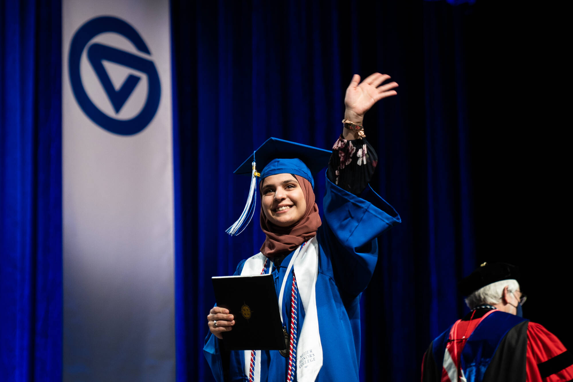An honors graduate walks across the stage at commencement and waves to the crowd while holding their diploma
